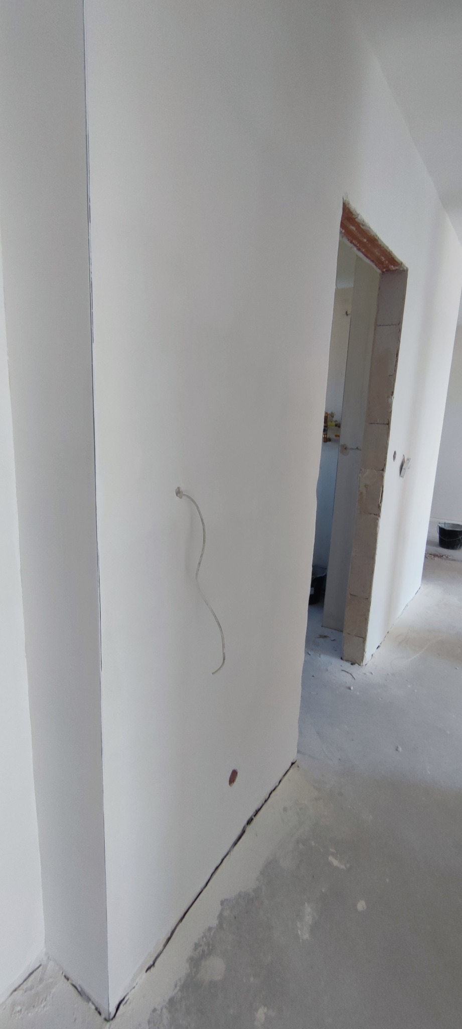 Biała ściana po szpachlowaniu z wystającym przewodem elektrycznym i widocznym wejściem do innego pomieszczenia w trakcie wykończenia.