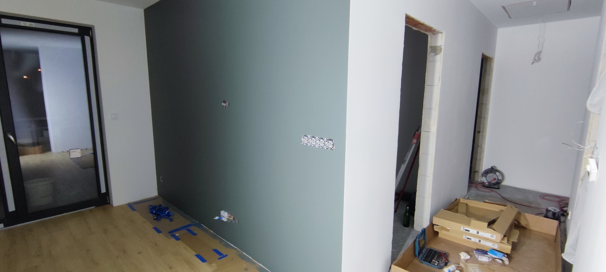Wnętrze w trakcie wykańczania: ściana pomalowana na szaro-zielono z gniazdkami elektrycznymi, podłoga z panelami. Widoczne narzędzia i kartony po materiałach.