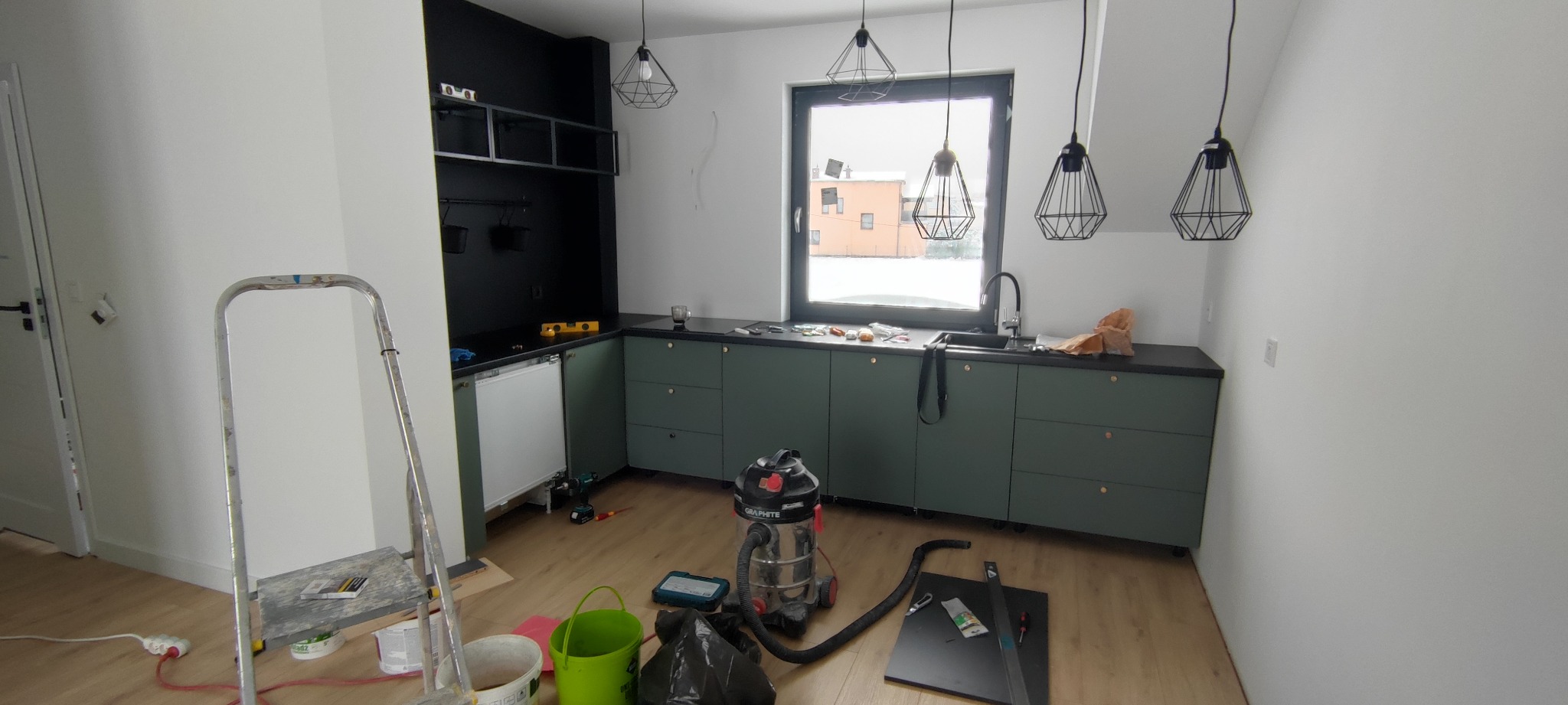 Montaż nowoczesnej kuchni z zielonymi szafkami i czarnym blatem. Widoczne narzędzia i drabina malarska. W tle okno i minimalistyczne lampy.