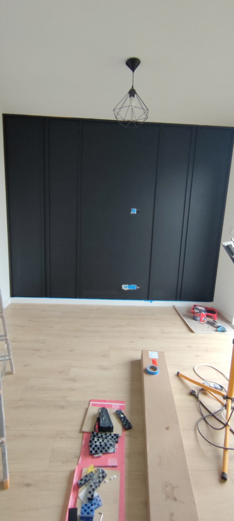 Ściana z czarnymi panelami dekoracyjnymi w trakcie montażu, widoczne gniazdka elektryczne, narzędzia i lampa sufitowa w minimalistycznym stylu.