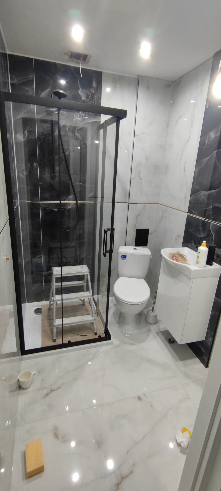 Wykończona łazienka z kabiną prysznicową typu walk-in z czarnymi ramami, białą armaturą i marmurowymi płytkami. Widoczna drabina malarska w kabinie.