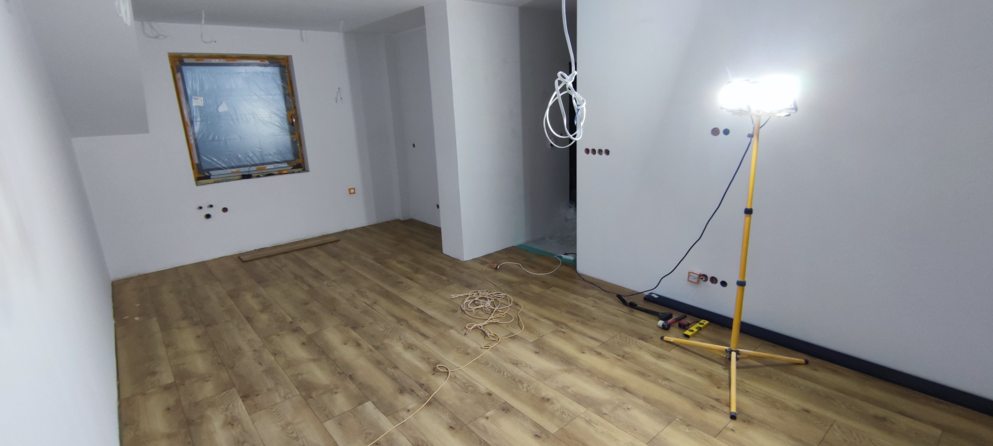 Układanie paneli podłogowych w jasnym wnętrzu. Widoczne narzędzia i lampa oświetlająca. Okno zabezpieczone folią. Częściowo wykończone pomieszczenie.