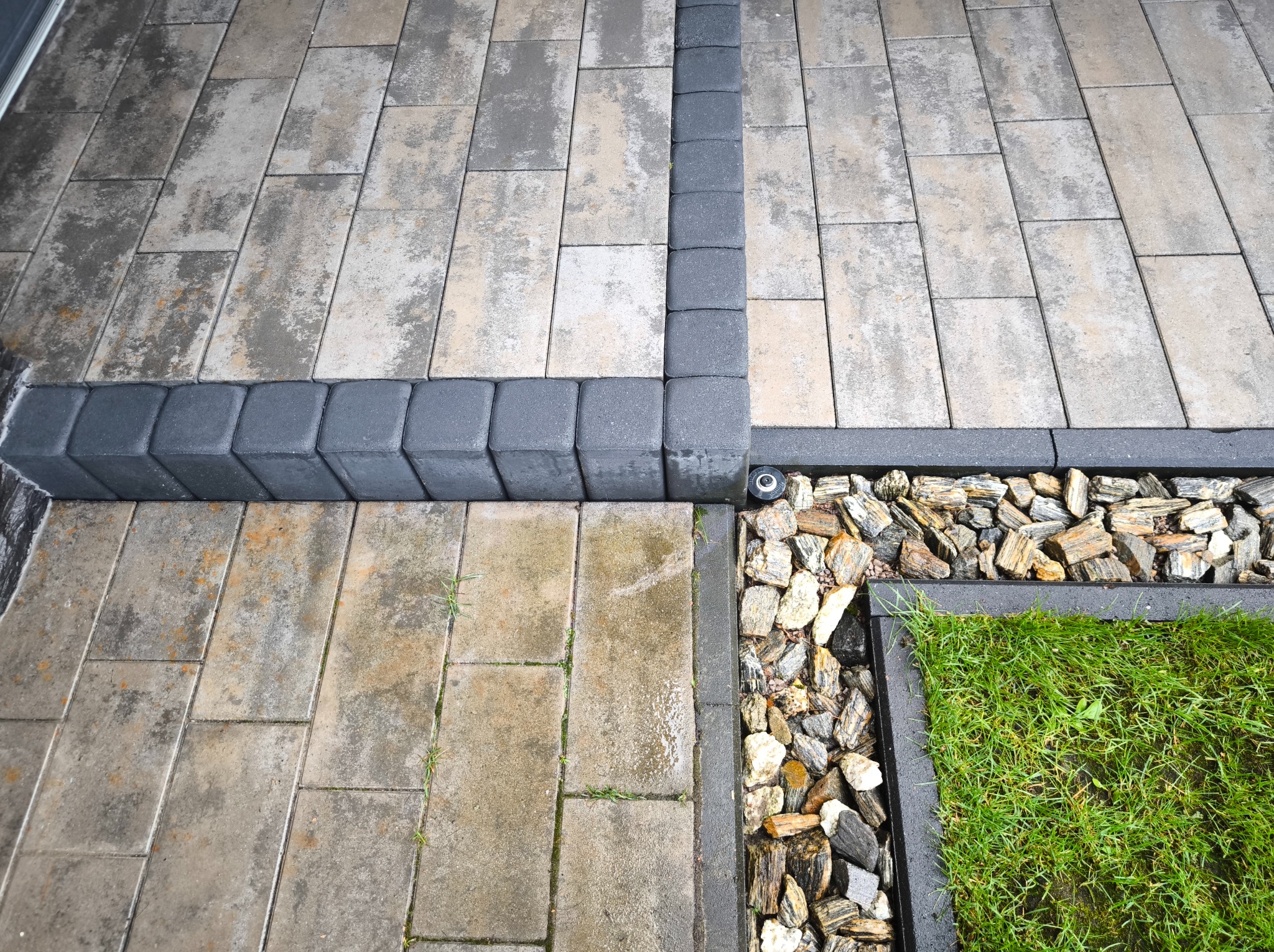 Ułożona kostka brukowa w szaro-beżowych odcieniach, obramowana ciemnoszarą kostką i ozdobnym kamieniem. Fragment trawnika w rogu. Widok z góry.