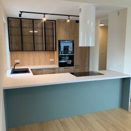 Prastora Dzianis Haponenka - Nowoczesna kuchnia z drewnianymi frontami, czarnymi akcentami i wyspą w odcieniu zieleni. Charakterystyczny okap cylindryczny i oświetlenie szynowe.