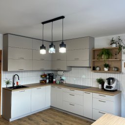 Prastora Dzianis Haponenka - Jasna kuchnia w kształcie litery L z drewnianym blatem, białymi szafkami z poziomymi uchwytami i trzema lampami wiszącymi nad blatem. Uwagę przykuwa drewniana półka z roślinami.