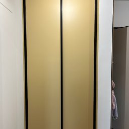 Prastora Dzianis Haponenka - Beżowa szafa wnękowa z przesuwnymi drzwiami i czarnymi ramami, wkomponowana w białe ściany, z widocznym fragmentem wnętrza szafy i drewnianą podłogą.