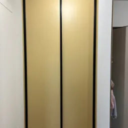 Beżowa szafa wnękowa z przesuwnymi drzwiami i czarnymi ramami, wkomponowana w białe ściany, z widocznym fragmentem wnętrza szafy i drewnianą podłogą.