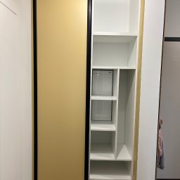Prastora Dzianis Haponenka - Wnętrze: szafa wnękowa z przesuwnymi drzwiami w kolorze złota, czarną ramą i białymi półkami. Widoczne dwa małe schowki z drzwiczkami. Nowoczesny design.