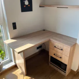 Narożne biurko z szufladami i półkami w jasnym drewnie, oświetlone promieniami słońca. Na ścianie wisi plakat z pingwinem.