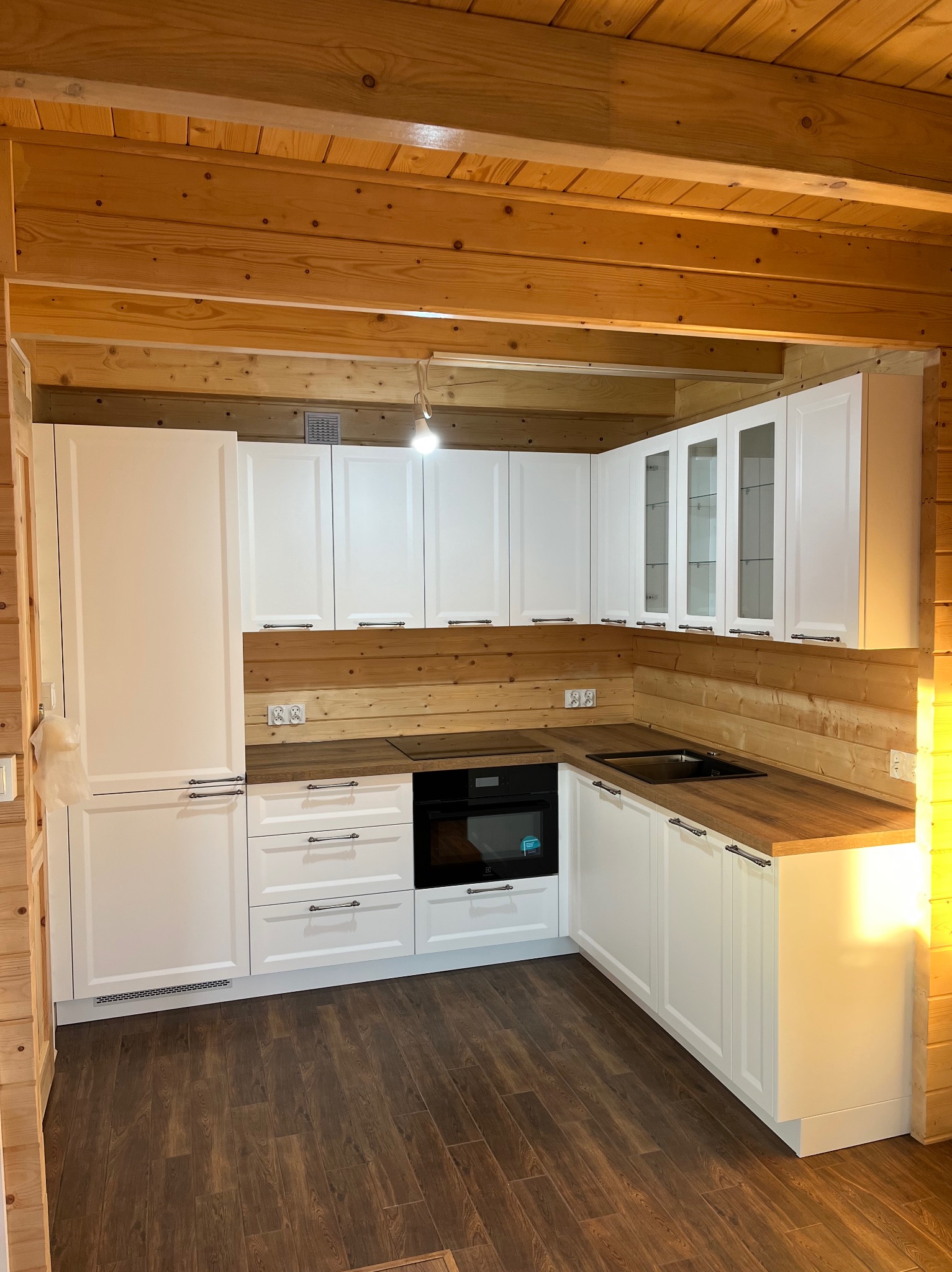 Biała kuchnia w domu z drewnianych bali: fronty szafek z frezowanymi ramkami, drewniany blat, czarny piekarnik i zlew. U góry szafki z przeszkleniami.