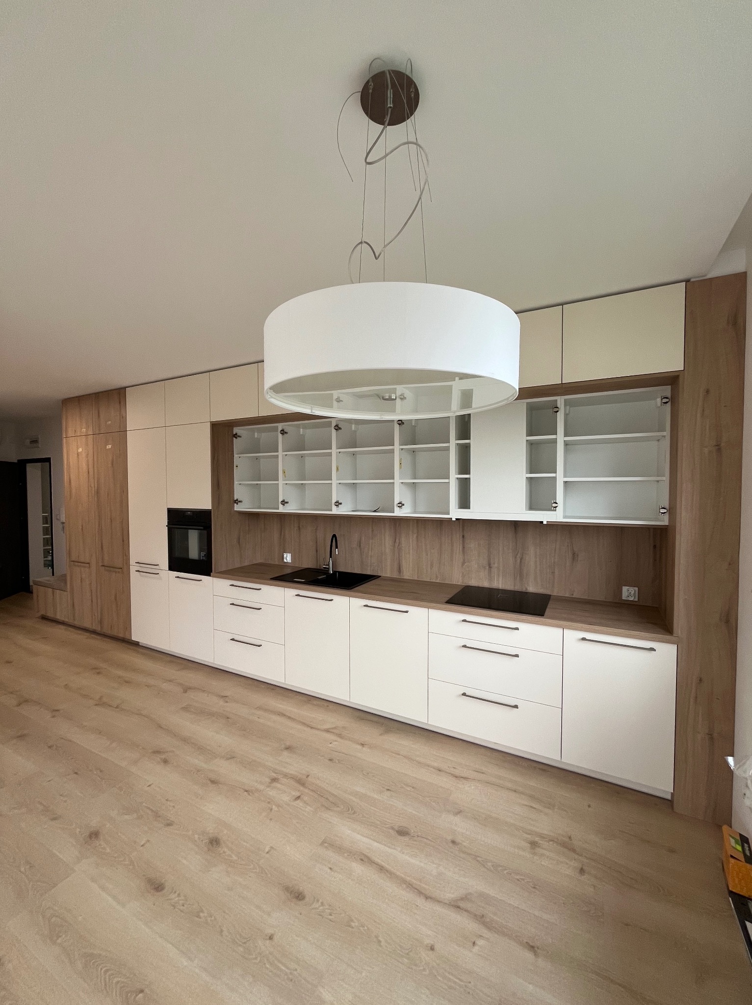 Nowoczesna kuchnia na wymiar z białymi frontami i drewnianymi akcentami. Otwarta szafka z półkami, czarny zlew i płyta indukcyjna. Jasne drewno na podłodze.