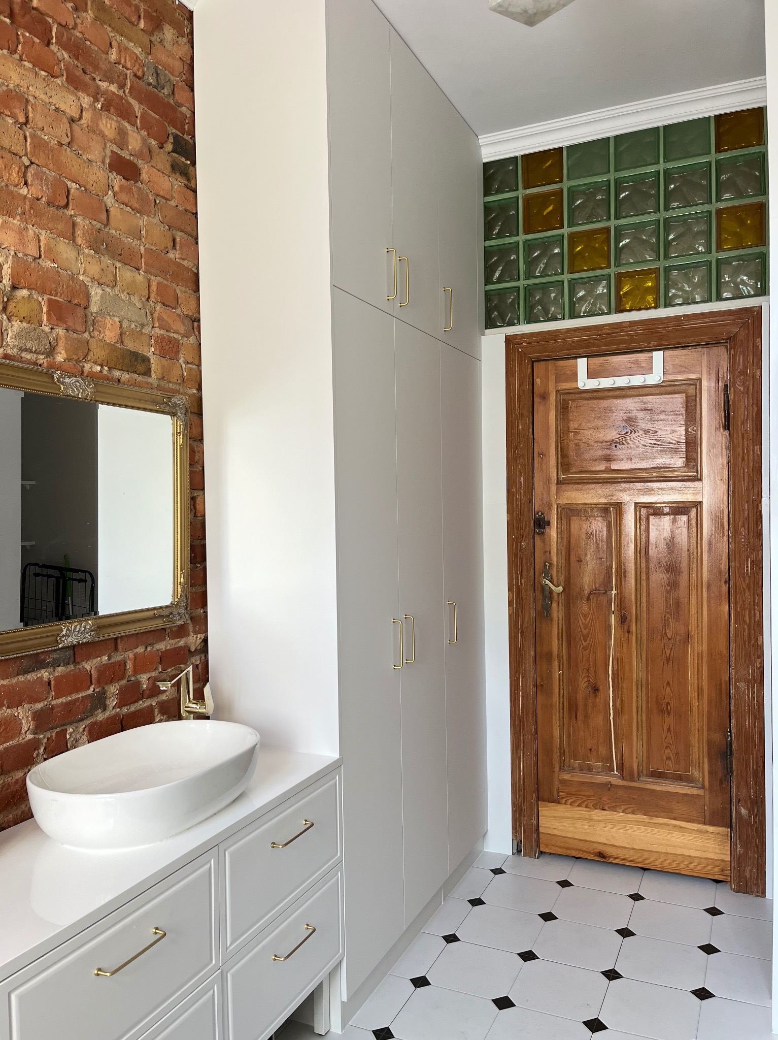 Nowoczesna łazienka z białą szafką pod umywalkę i wysoką szafą w zabudowie, złote uchwyty, ceglana ściana i lustro w złotej ramie. Kontrastowe płytki podłogowe.