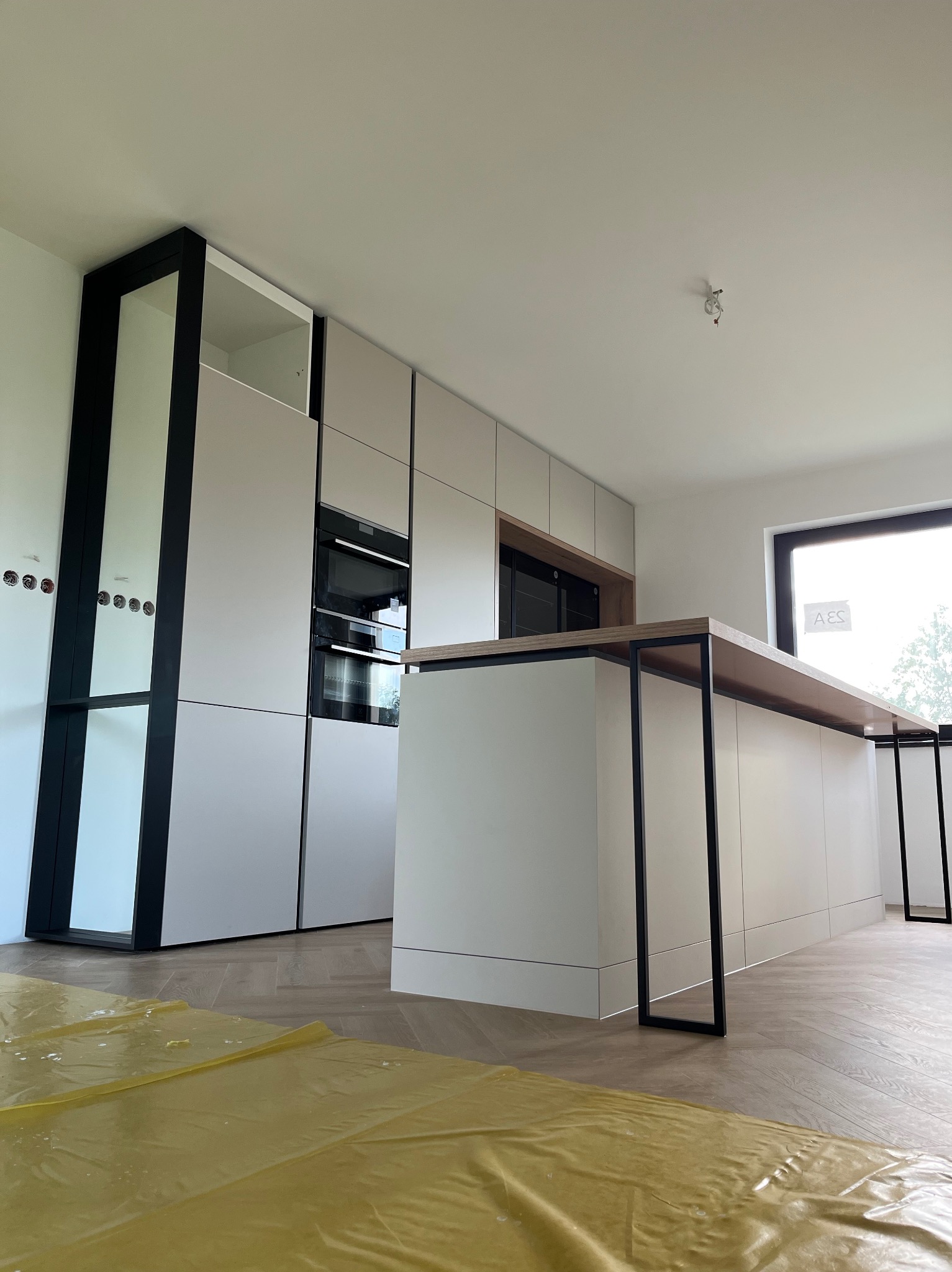 Nowoczesna kuchnia z wyspą: białe fronty, czarne ramy i drewniany blat. Podłoga zabezpieczona folią, widoczne gniazdka i okno. Minimalistyczny design.