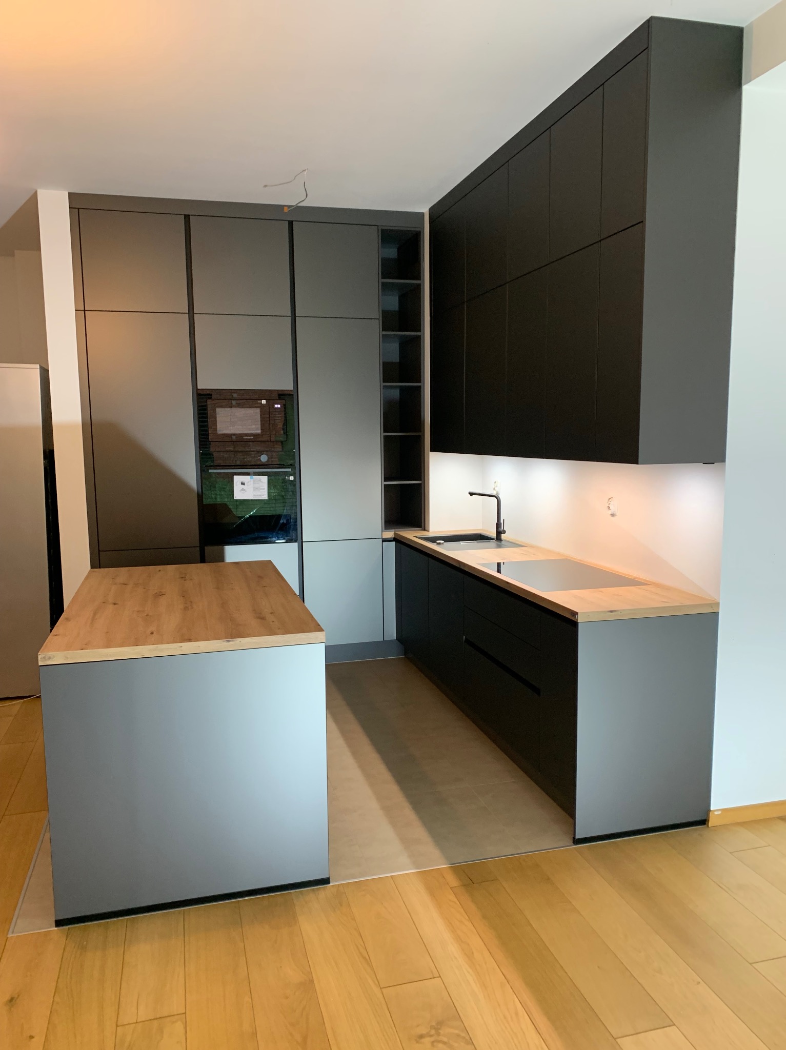 Nowoczesna kuchnia z wyspą: szare i czarne szafki, drewniany blat, oświetlenie LED pod szafkami. Uwagę przykuwa nowoczesny design i minimalistyczne wykończenie.