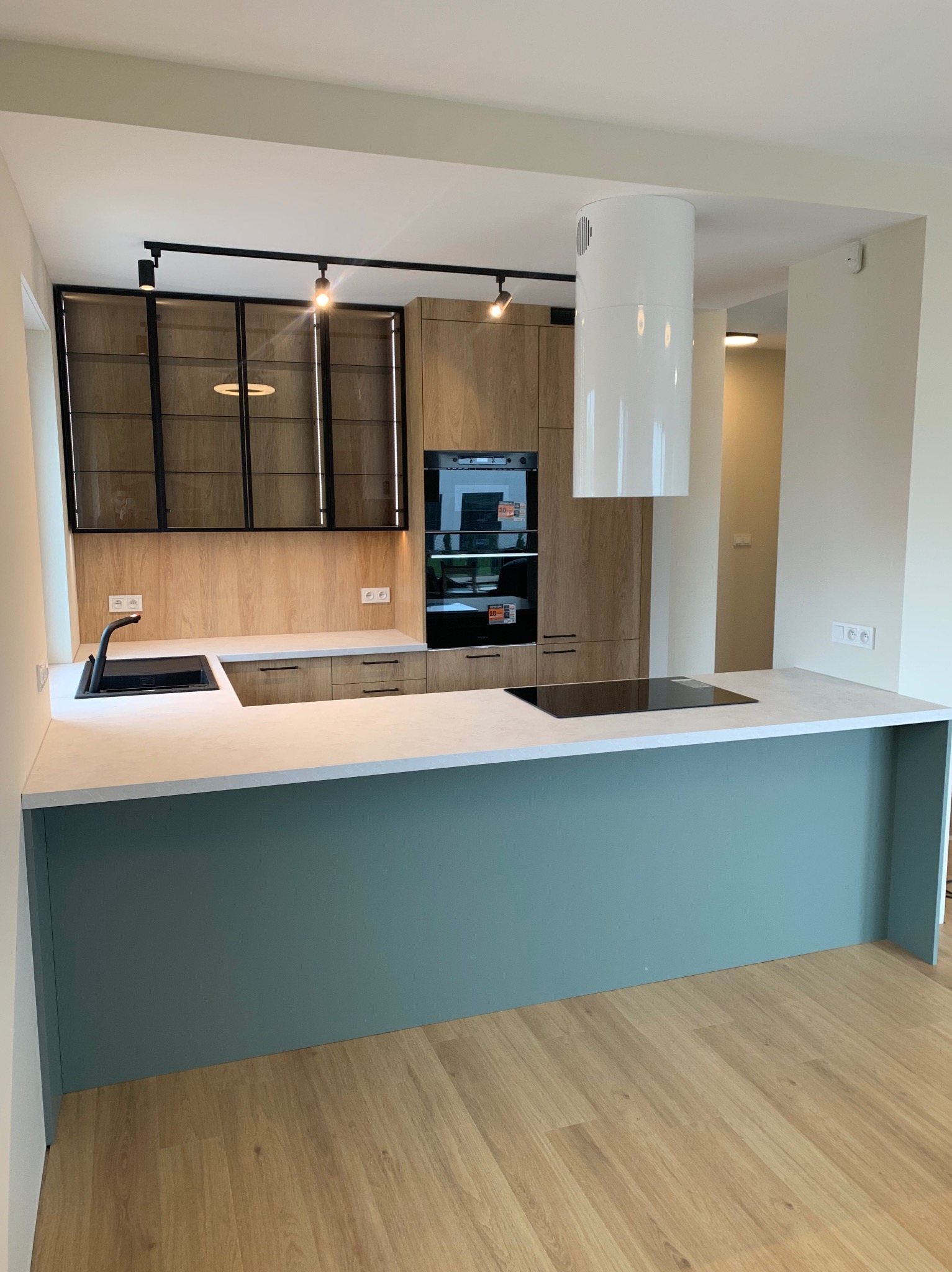 Nowoczesna kuchnia z drewnianymi frontami, czarnymi akcentami i wyspą w odcieniu zieleni. Charakterystyczny okap cylindryczny i oświetlenie szynowe.