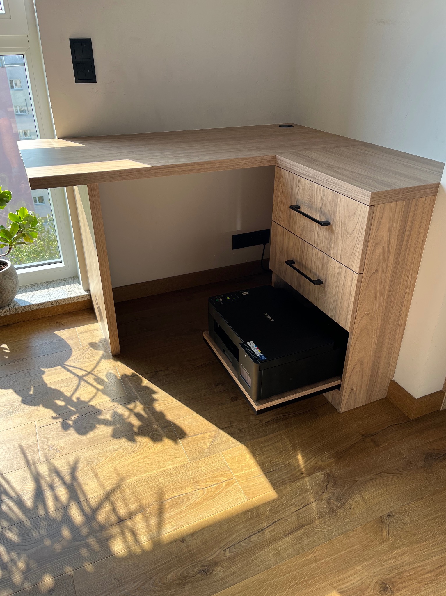 Biurko narożne z szufladami i wnęką na drukarkę, wykonane z drewna w odcieniach brązu. Na podłodze widoczne cienie roślin. Nowoczesny design.