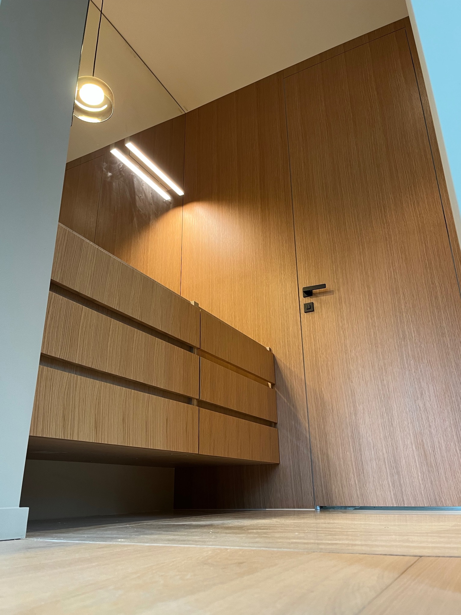 Drewniana zabudowa: szafa z ukrytymi drzwiami i komoda z szufladami, oświetlenie LED, minimalistyczny styl. Ujęcie z dołu podkreśla wysokość zabudowy.