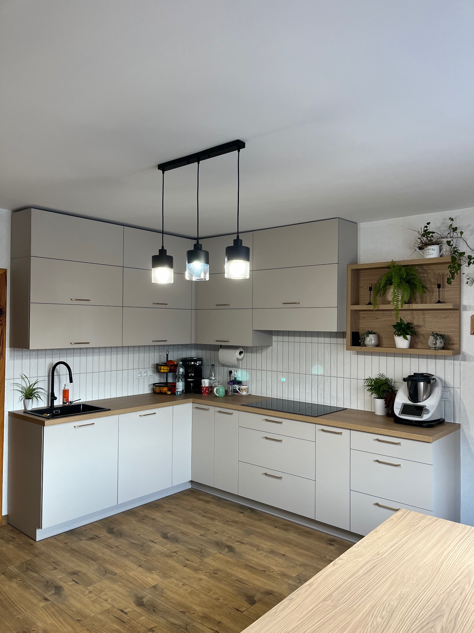 Jasna kuchnia w kształcie litery L z drewnianym blatem, białymi szafkami z poziomymi uchwytami i trzema lampami wiszącymi nad blatem. Uwagę przykuwa drewniana półka z roślinami.