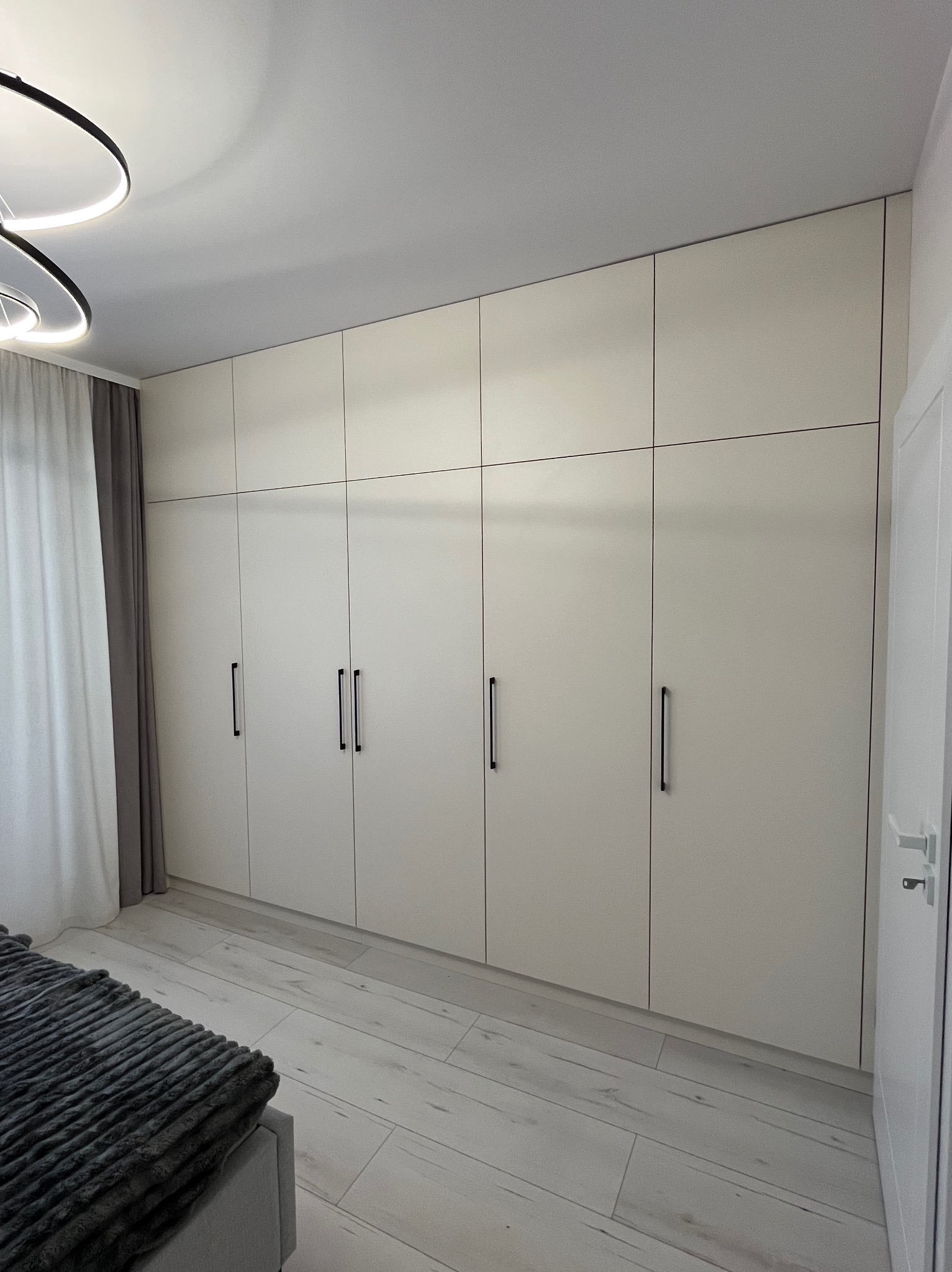 Duża, jasna szafa wnękowa z czarnymi uchwytami, zajmująca całą ścianę w sypialni. Widoczne panele podłogowe i fragment łóżka z narzutą.
