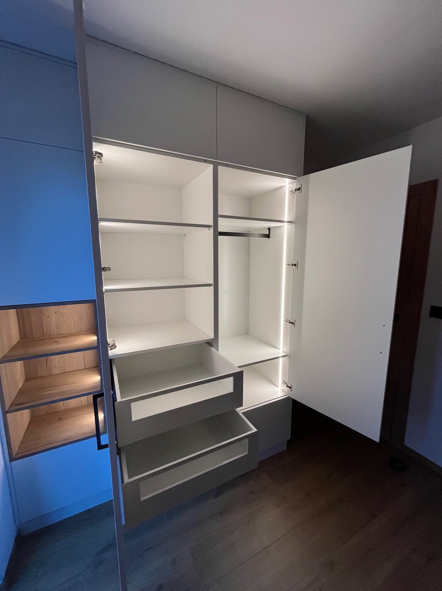 Otwarta szafa wnękowa z szufladami i półkami, oświetlona ledami. Jedna strona szafy w kolorze niebieskim z drewnianymi półkami.