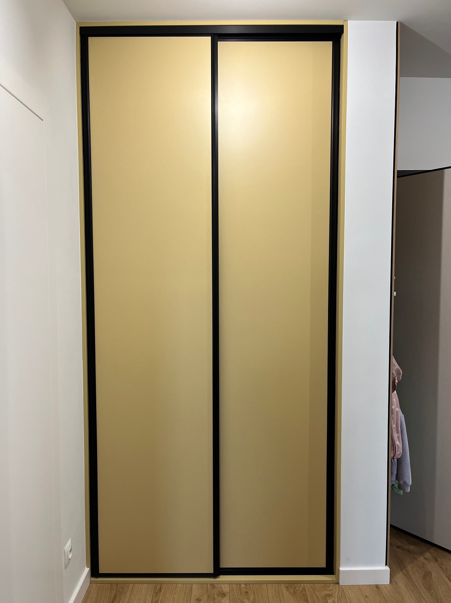 Beżowa szafa wnękowa z przesuwnymi drzwiami i czarnymi ramami, wkomponowana w białe ściany, z widocznym fragmentem wnętrza szafy i drewnianą podłogą.