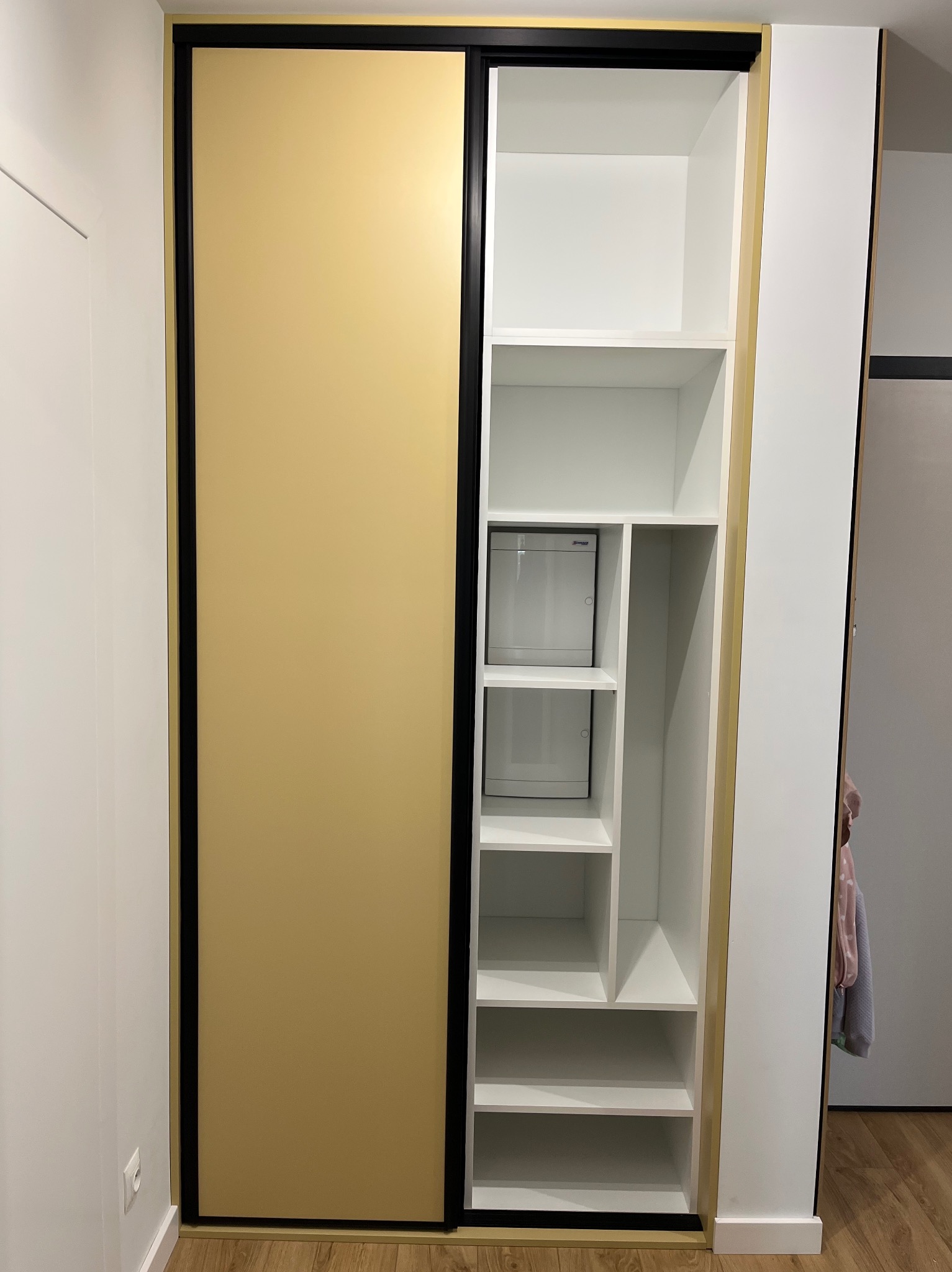 Wnętrze: szafa wnękowa z przesuwnymi drzwiami w kolorze złota, czarną ramą i białymi półkami. Widoczne dwa małe schowki z drzwiczkami. Nowoczesny design.