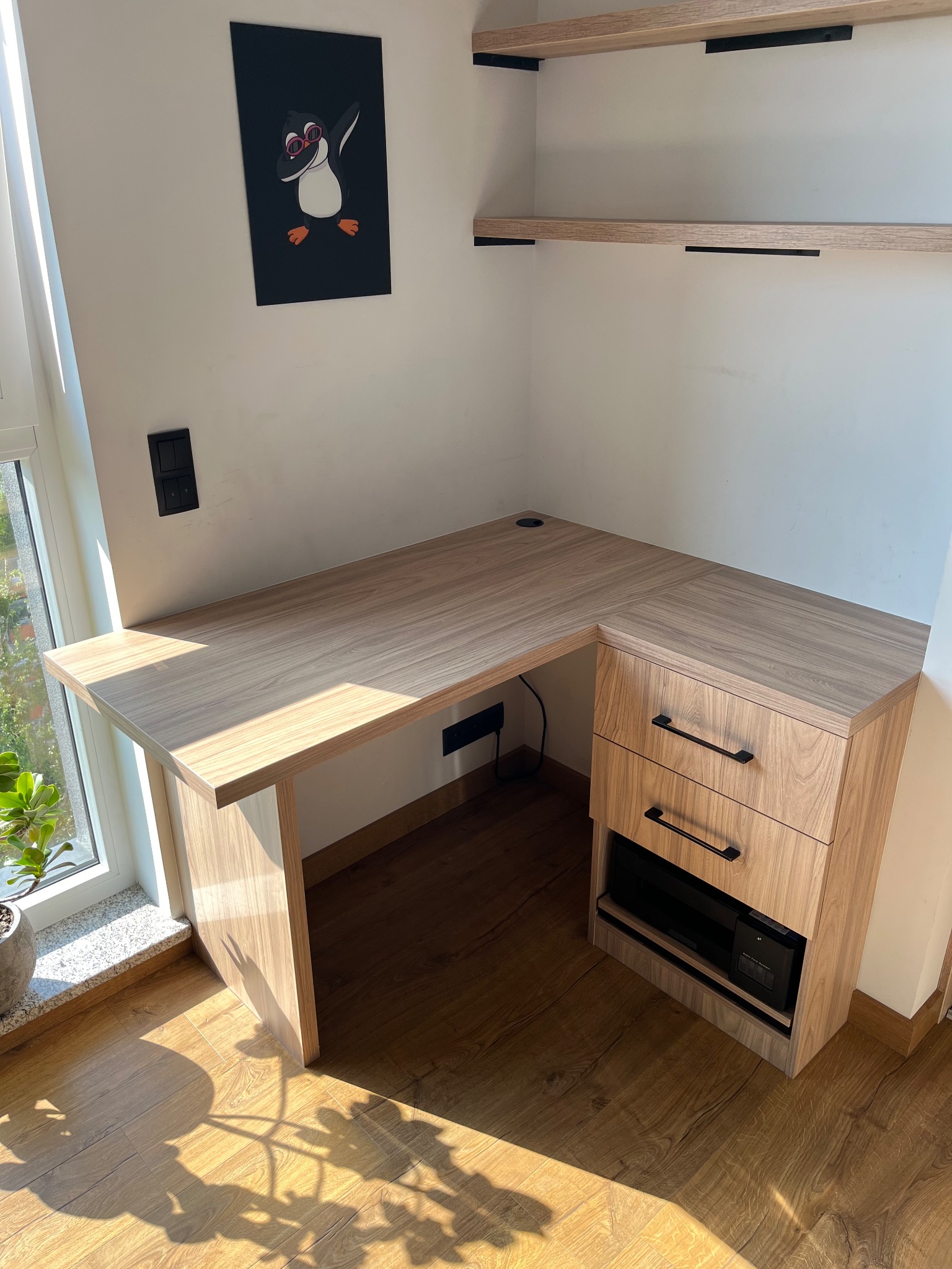 Narożne biurko z szufladami i półkami w jasnym drewnie, oświetlone promieniami słońca. Na ścianie wisi plakat z pingwinem.