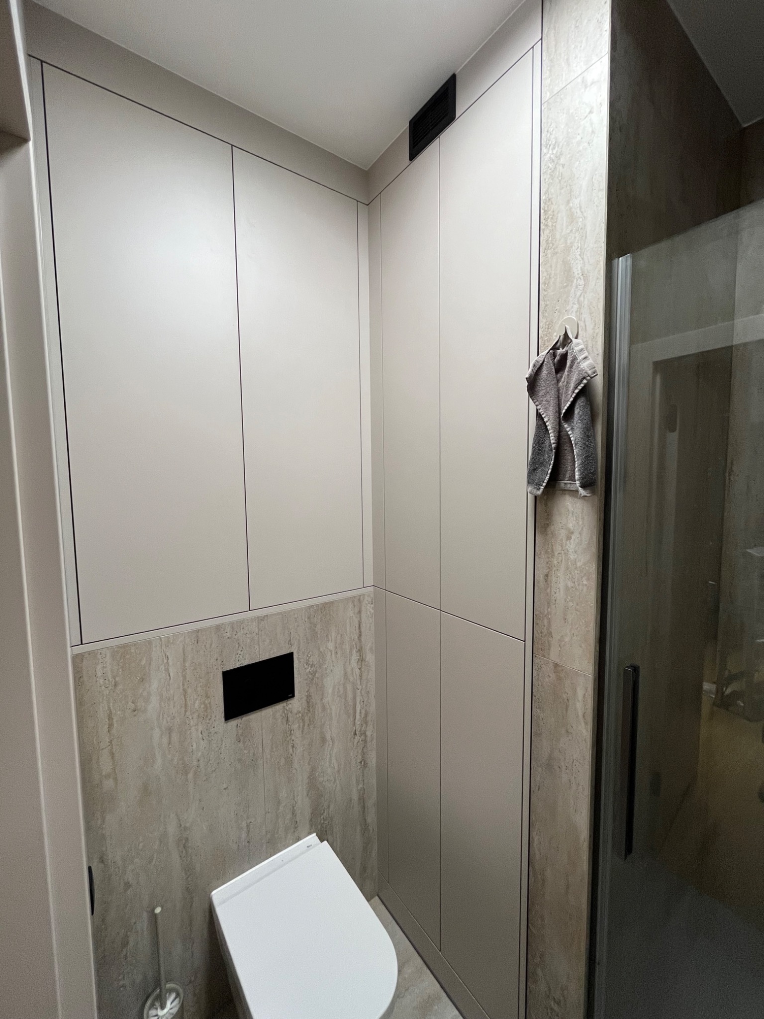 Jasna łazienka z zabudową meblową na wymiar, beżowe szafki od podłogi do sufitu, toaleta podwieszana, ręcznik na ścianie z kamienia.