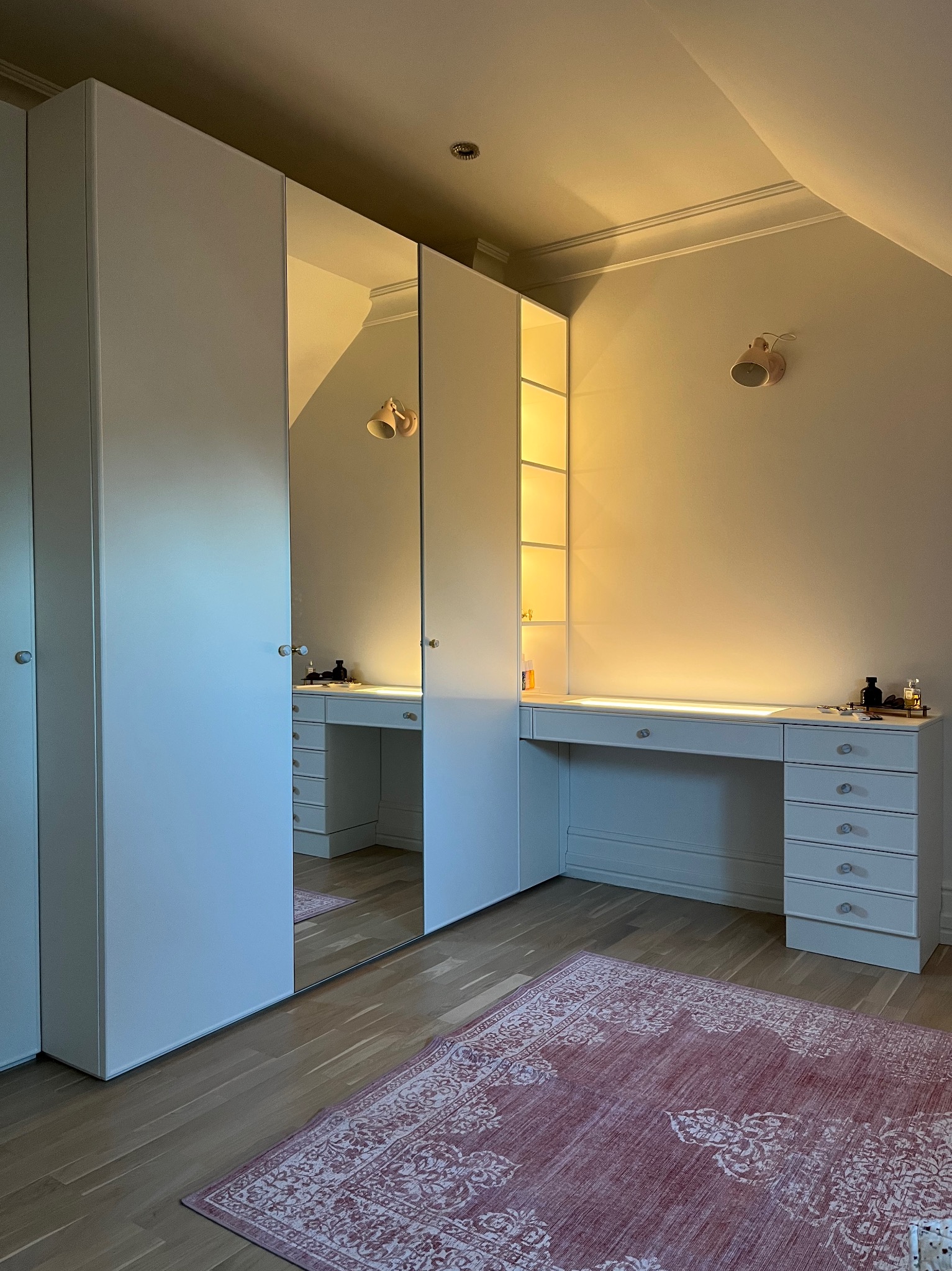 Biała szafa wnękowa z lustrem i toaletką w pokoju na poddaszu, oświetlenie punktowe, dywan w orientalny wzór. Widok na gotową realizację.