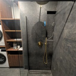 PORTO BAU OLEKSANDR ROMANETSKYI - Nowoczesna kabina prysznicowa z deszczownicą w kolorze złotym na tle ciemnych, kamiennych płytek. Widoczny odpływ liniowy i szklana ścianka kabiny.