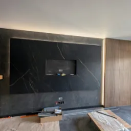 Wykończona ściana z czarnych płytek z złotymi żyłami i wnęką na TV, podświetlona listwami LED. Obok drewniana ściana lamelowa. W tle widoczne gniazdka elektryczne.