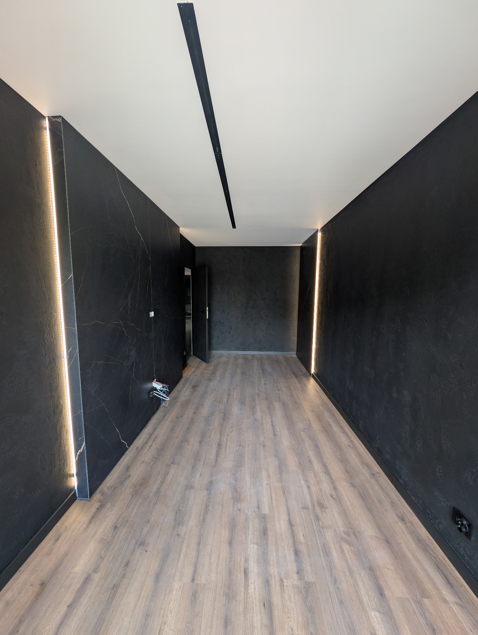 Nowoczesne wykończenie wnętrza: ciemne ściany z efektem marmuru, podświetlenie LED w pionowych listwach, drewniana podłoga i minimalistyczny sufit z czarną listwą.