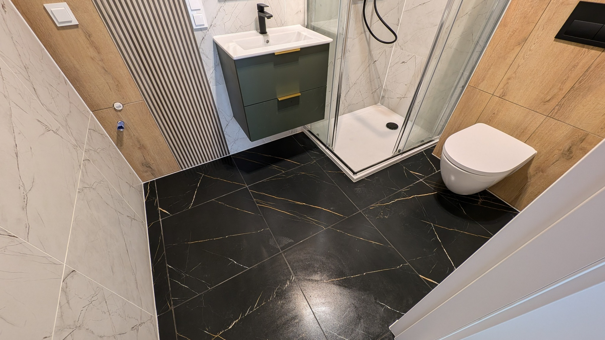 Nowoczesna łazienka z czarną, marmurkową podłogą, kabiną prysznicową i wiszącą szafką umywalkową w odcieniach zieleni, wykończona płytkami imitującymi drewno.
