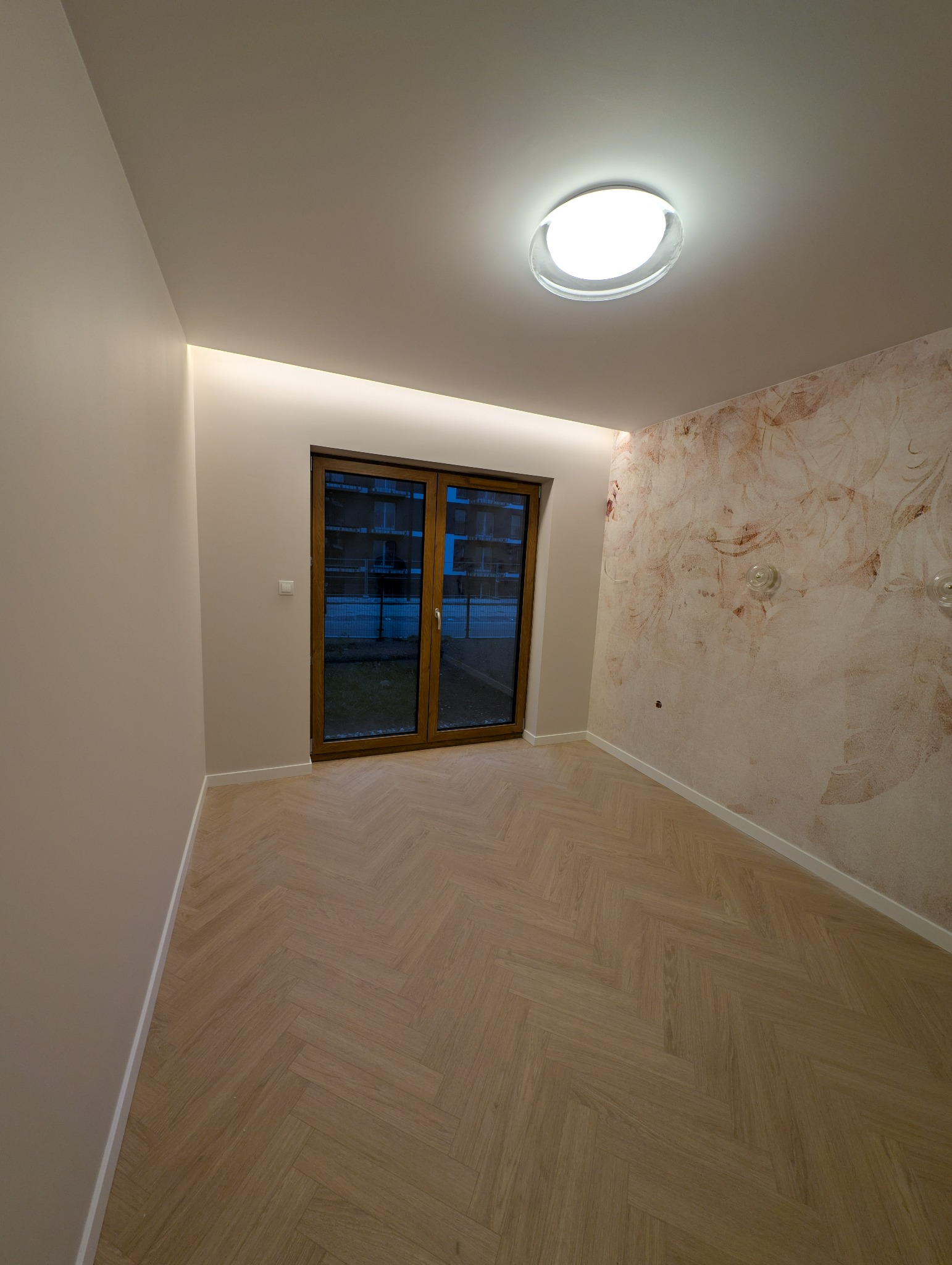 Jasny pokój po remoncie z parkietem w jodełkę, beżowymi ścianami i wzorzystą tapetą na jednej ścianie. Widoczne okno balkonowe i nowoczesna lampa sufitowa.