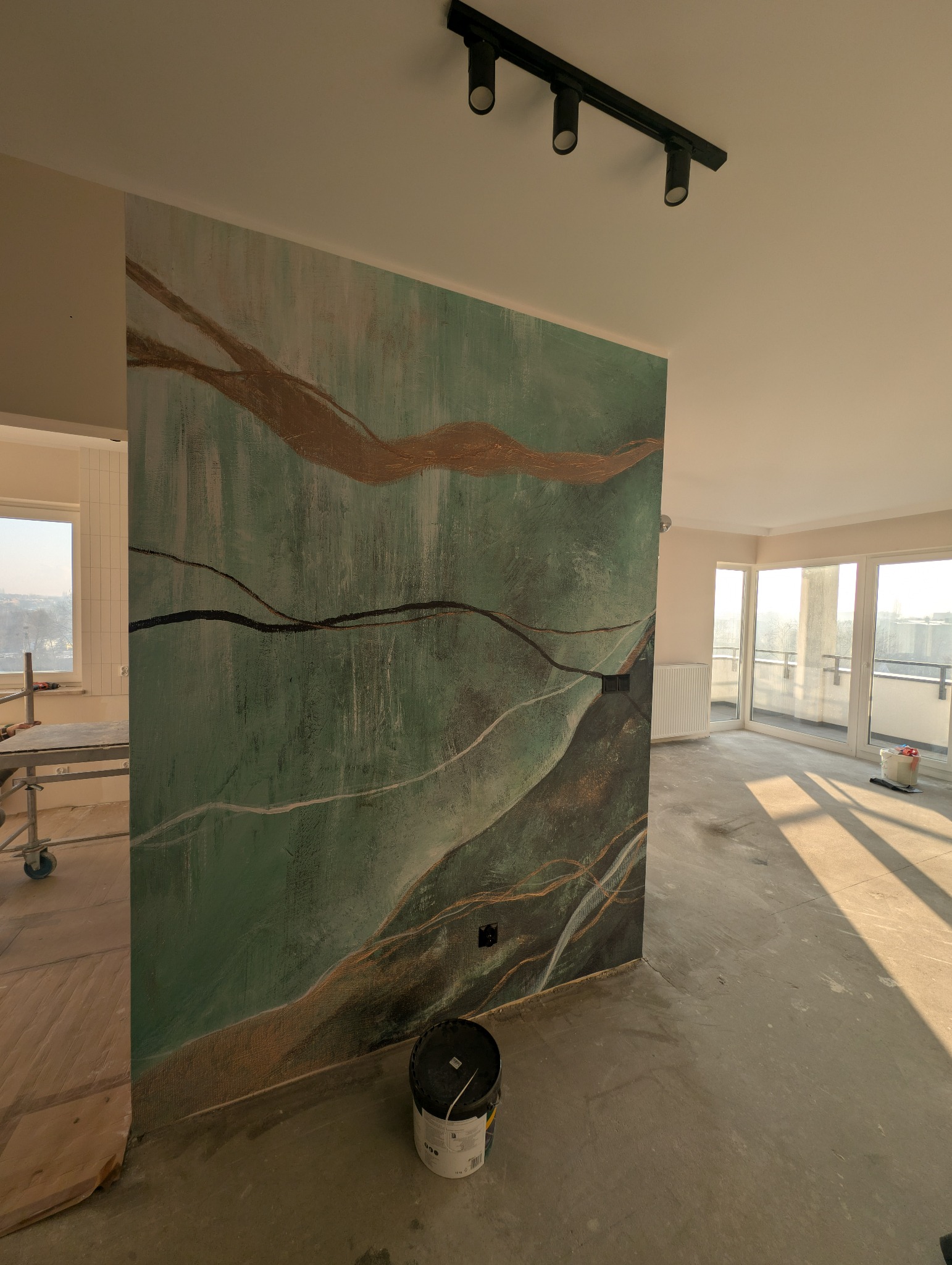 Ściana z dekoracyjną tapetą w odcieniach zieleni i złota w Gliwicach, oświetlona reflektorami. Widoczny fragment rusztowania i wiadro z farbą.