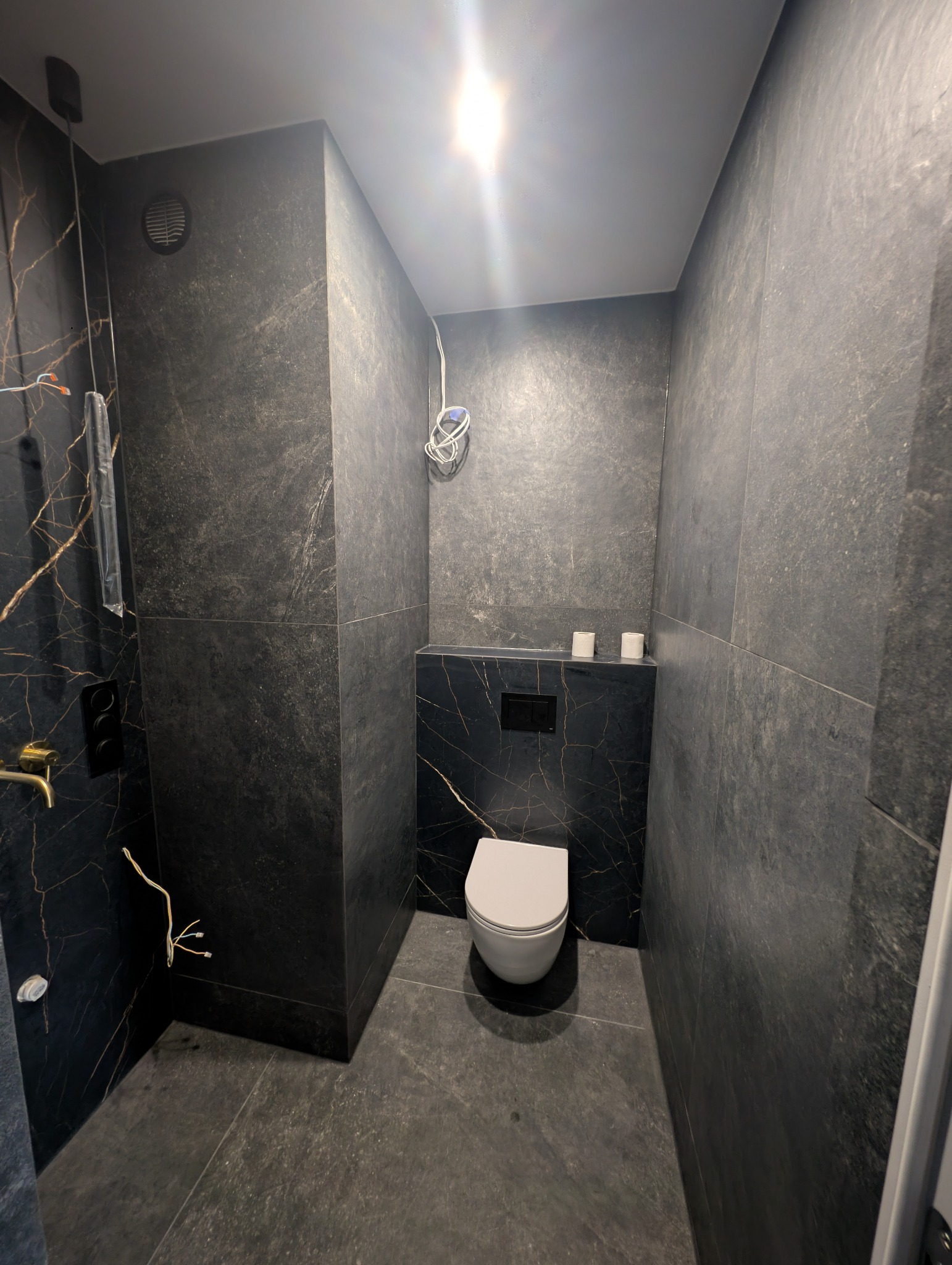 Nowoczesna toaleta z czarnymi płytkami imitującymi marmur ze złotymi żyłami. Widoczne przyłącza elektryczne i hydrauliczne w trakcie montażu.