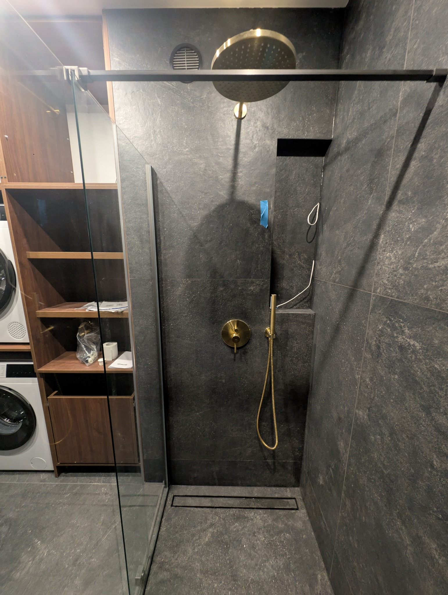 Nowoczesna kabina prysznicowa z deszczownicą w kolorze złotym na tle ciemnych, kamiennych płytek. Widoczny odpływ liniowy i szklana ścianka kabiny.