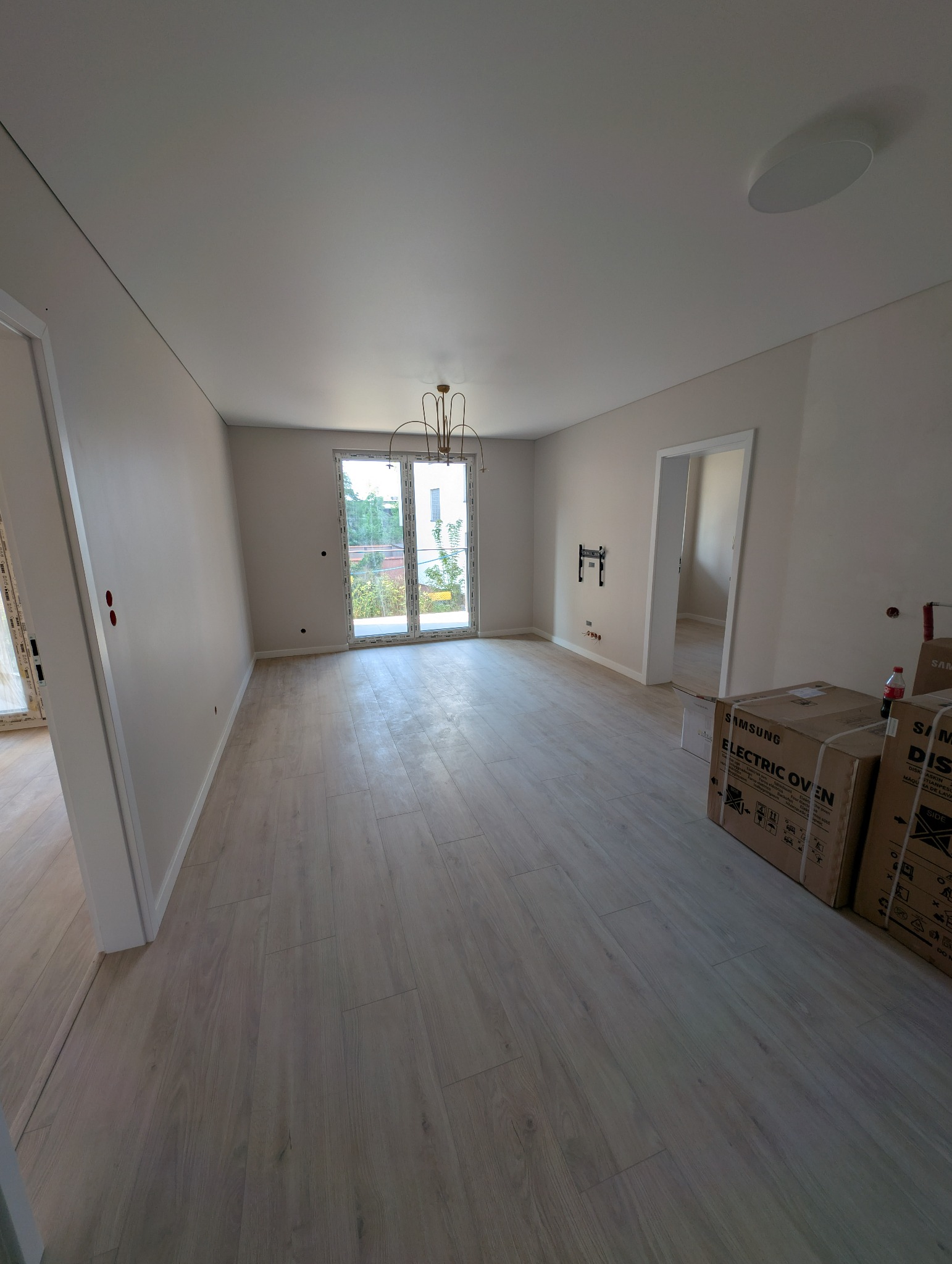 Jasne wnętrze po remoncie: panele podłogowe, świeżo pomalowane ściany w odcieniach beżu,  okno balkonowe z widokiem na zieleń, kartony z nowym sprzętem.