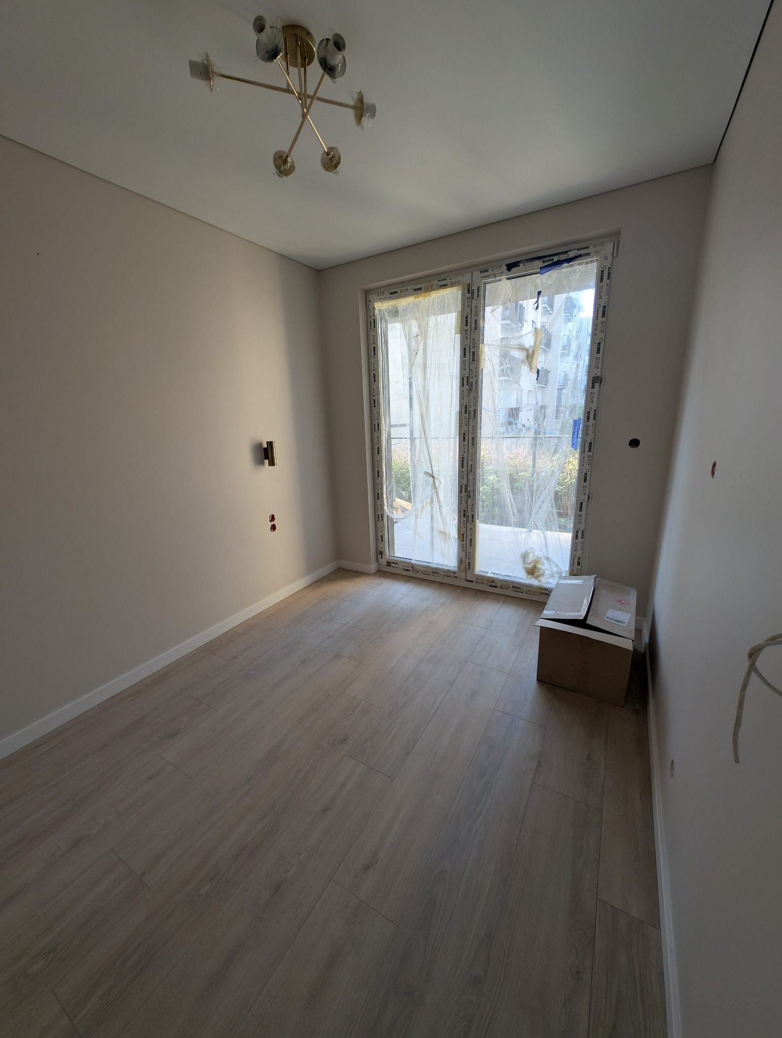 Jasne panele podłogowe w pokoju po remoncie, widoczne okno z zasłoniętą folią, karton i minimalistyczna lampa sufitowa. Ściany pomalowane na jasny kolor.