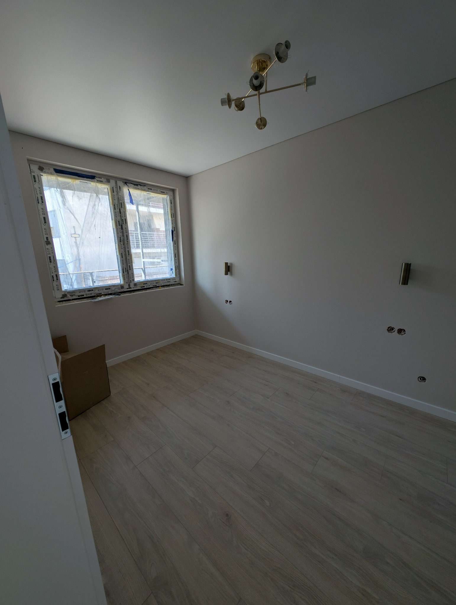 Jasne wnętrze pokoju po remoncie, z panelami podłogowymi, oknem z widokiem na zewnątrz i minimalistycznym oświetleniem sufitowym. Widoczne gniazdka elektryczne.