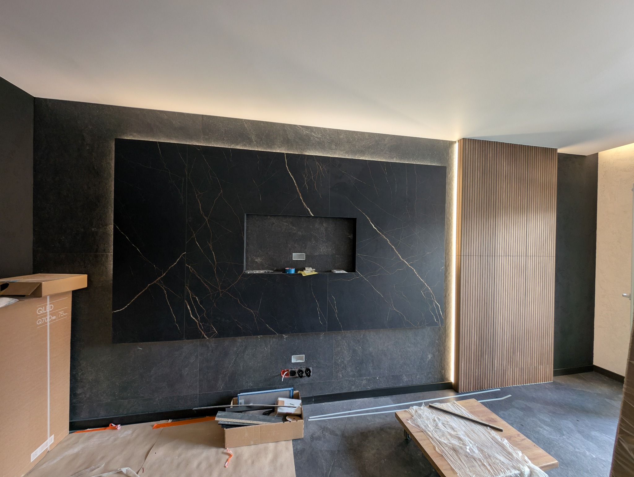 Wykończona ściana z czarnych płytek z złotymi żyłami i wnęką na TV, podświetlona listwami LED. Obok drewniana ściana lamelowa. W tle widoczne gniazdka elektryczne.