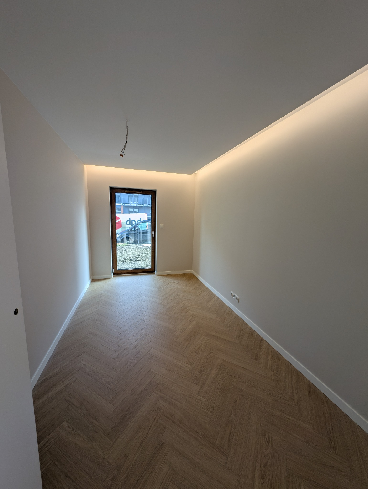 Pusty pokój z parkietem w jodełkę, świeżo pomalowanymi ścianami i oświetleniem LED przy suficie. Widoczne okno/drzwi balkonowe i wystające kable.