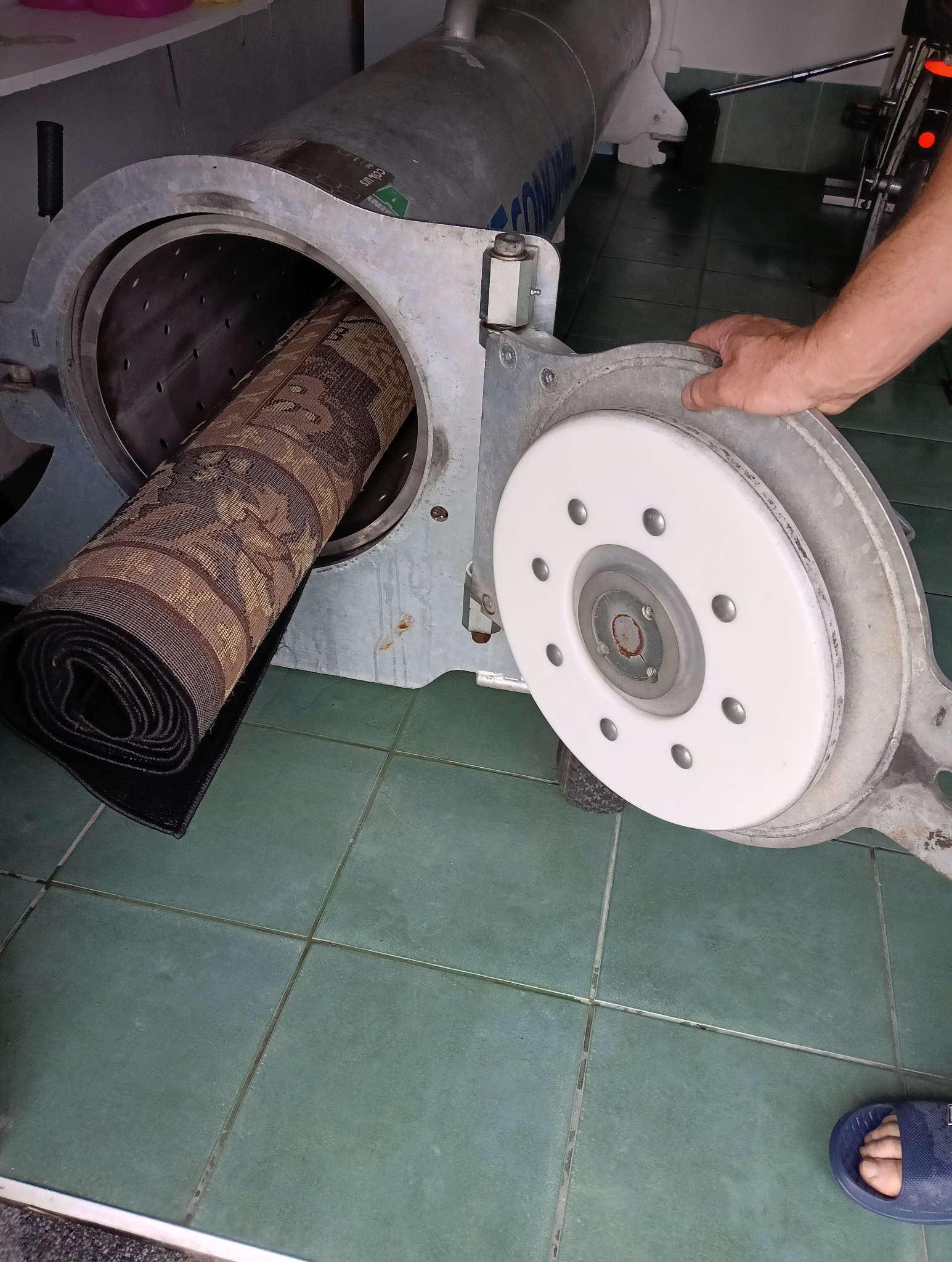 Dywan zwinięty w rolkę, umieszczony w przemysłowej wirówce do prania dywanów. Otwarta klapa urządzenia, zielona podłoga z płytek.