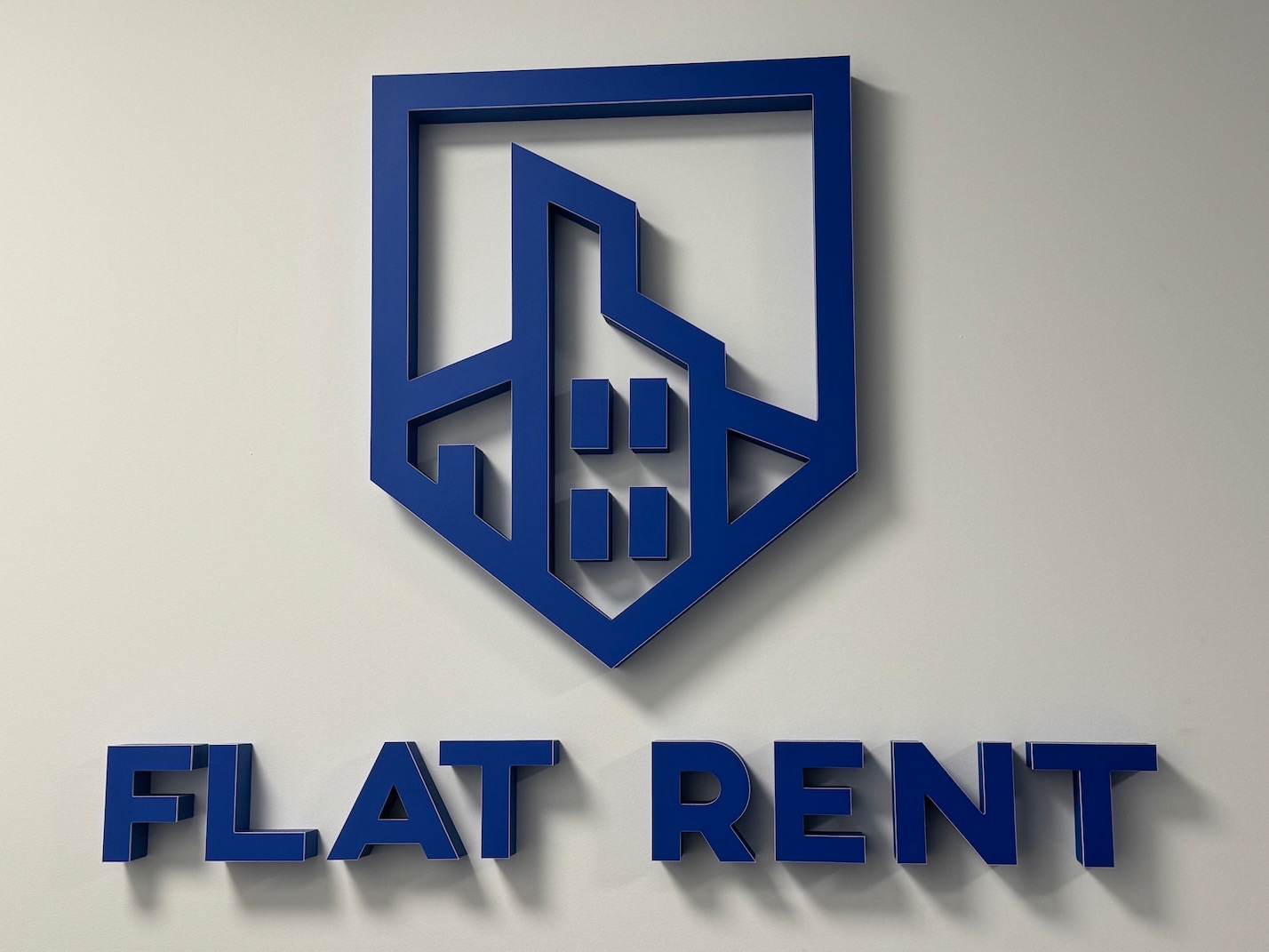 Niebieskie logo 'FLAT RENT' na białej ścianie. Geometryczny symbol domu w górnej części. Minimalistyczny design, ostre cienie.