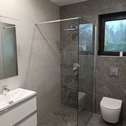 Roman Halets - Nowoczesna łazienka z szarymi płytkami imitującymi kamień, kabiną prysznicową typu walk-in, podwieszaną toaletą i umywalką z szafką. Widok na okno z zielenią na zewnątrz.