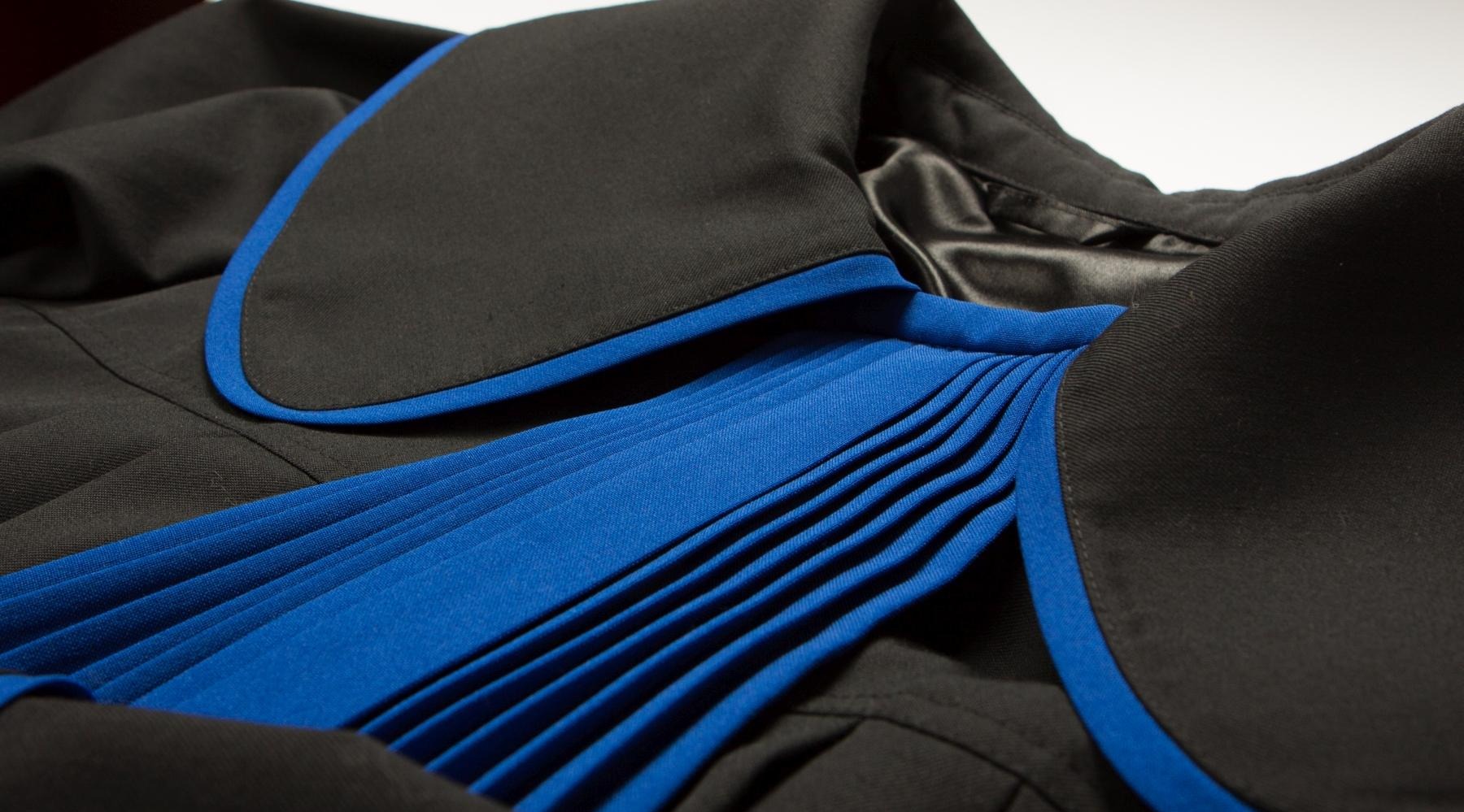 Zbliżenie na togę prawniczą: czarny materiał z niebieskimi plisami, eleganckie wykończenie. Detal stroju urzędowego w neutralnym oświetleniu.