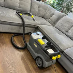 Szaro-żółty odkurzacz piorący Kärcher na drewnianej podłodze, obok szarej, welurowej sofy narożnej. W tle widoczne rośliny doniczkowe.