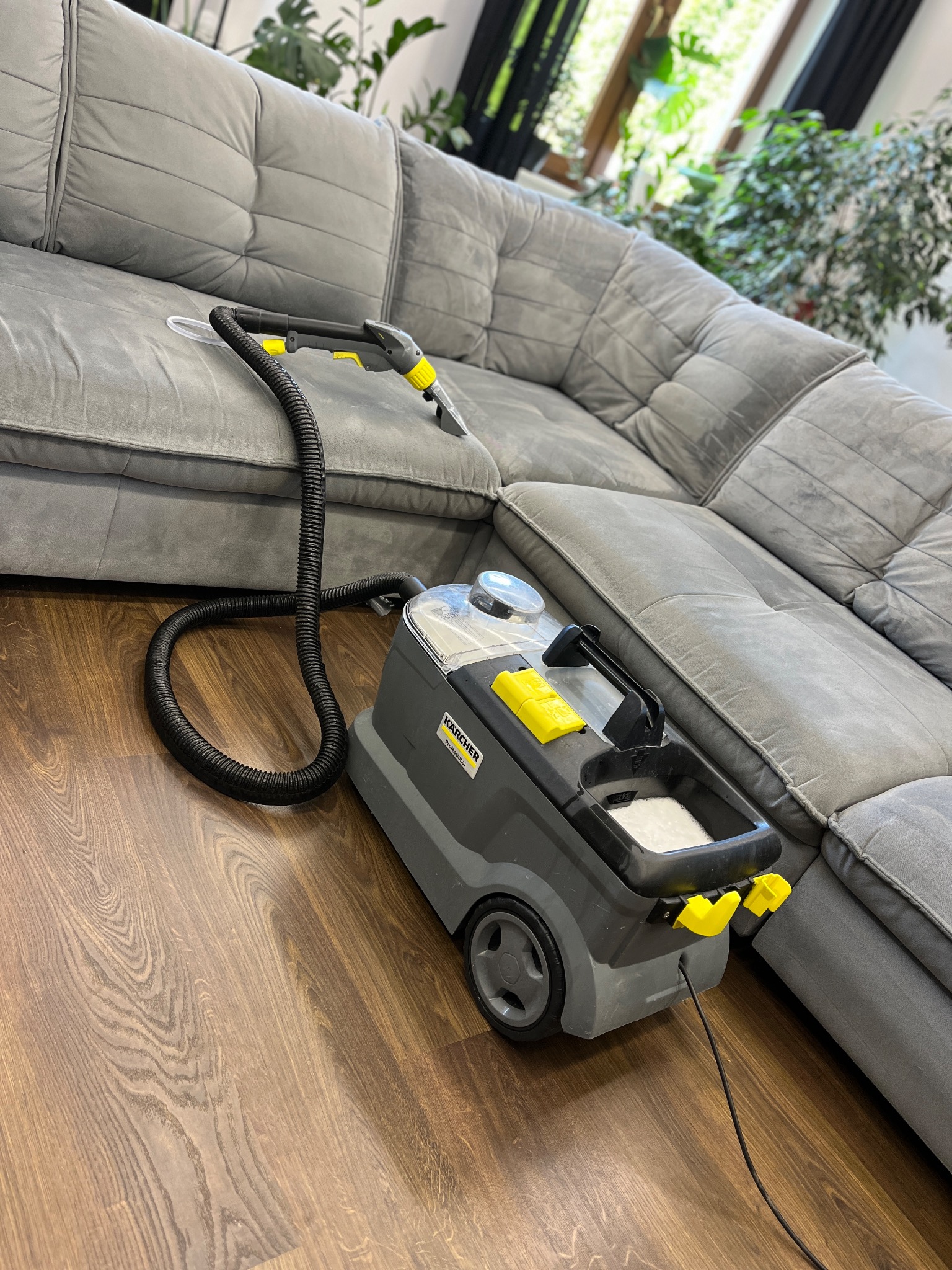 Szaro-żółty odkurzacz piorący Kärcher na drewnianej podłodze, obok szarej, welurowej sofy narożnej. W tle widoczne rośliny doniczkowe.