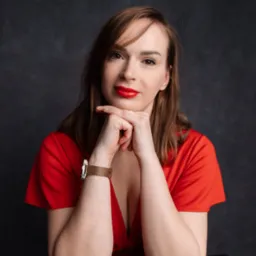 Portret kobiety w czerwonej sukience, z rękami pod brodą, patrzącej w obiektyw, z widocznym zegarkiem na ręce i ciemnym, teksturowanym tłem.