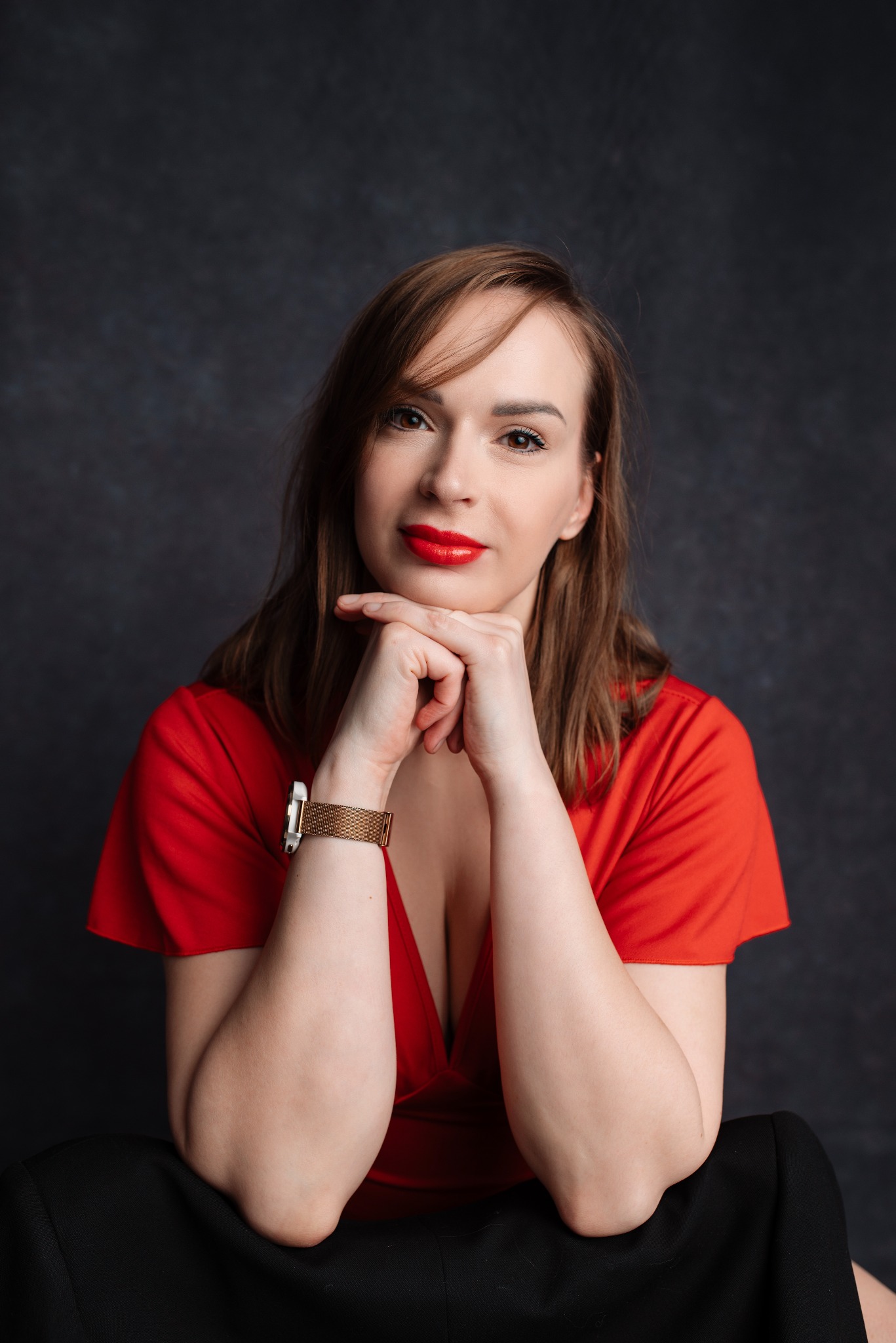 Portret kobiety w czerwonej sukience, z rękami pod brodą, patrzącej w obiektyw, z widocznym zegarkiem na ręce i ciemnym, teksturowanym tłem.